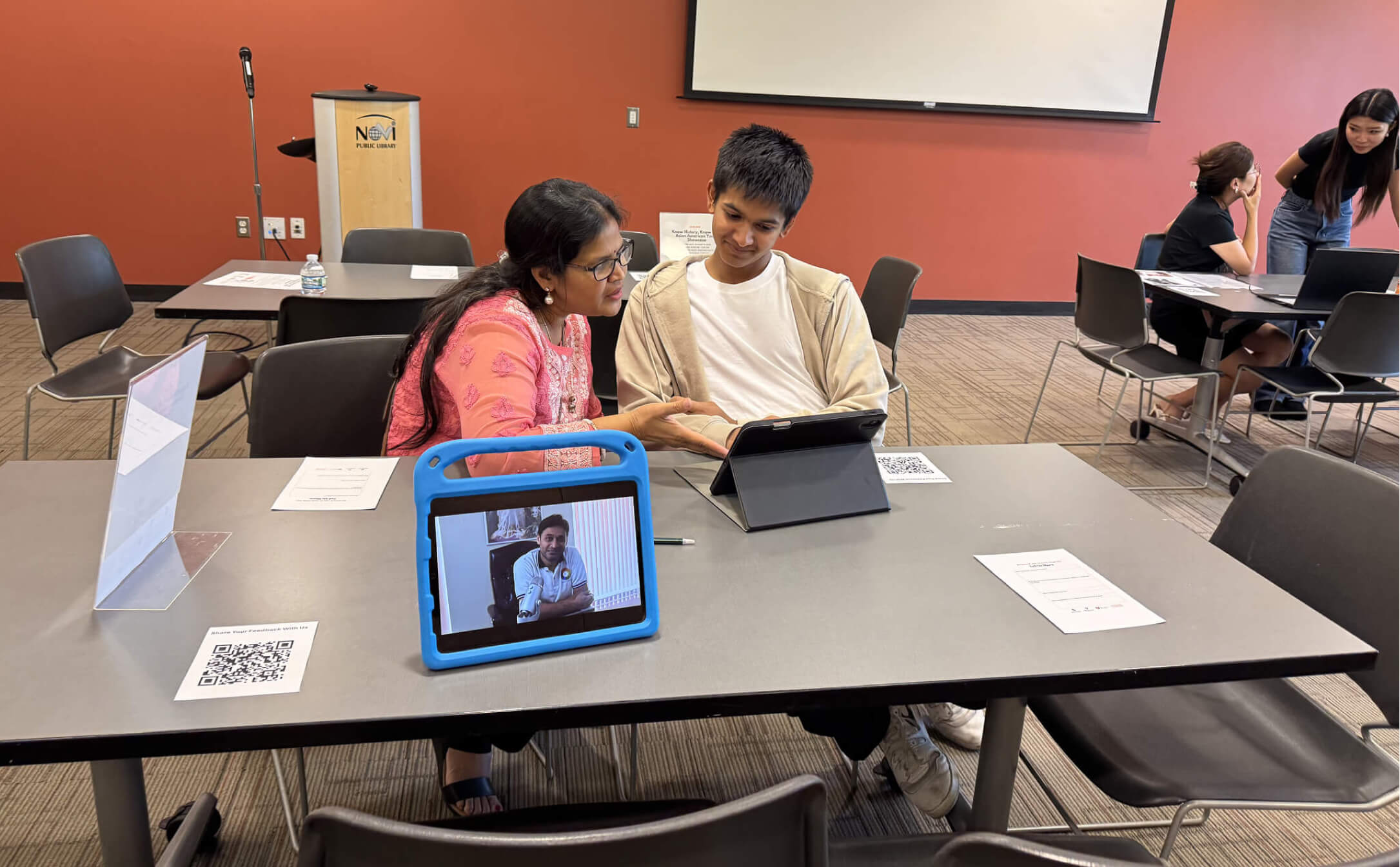 A woman in a pink outfit and a teenage boy sitting at a table in a library room, looking at a tablet, with another tablet showing a man's video call nearby.