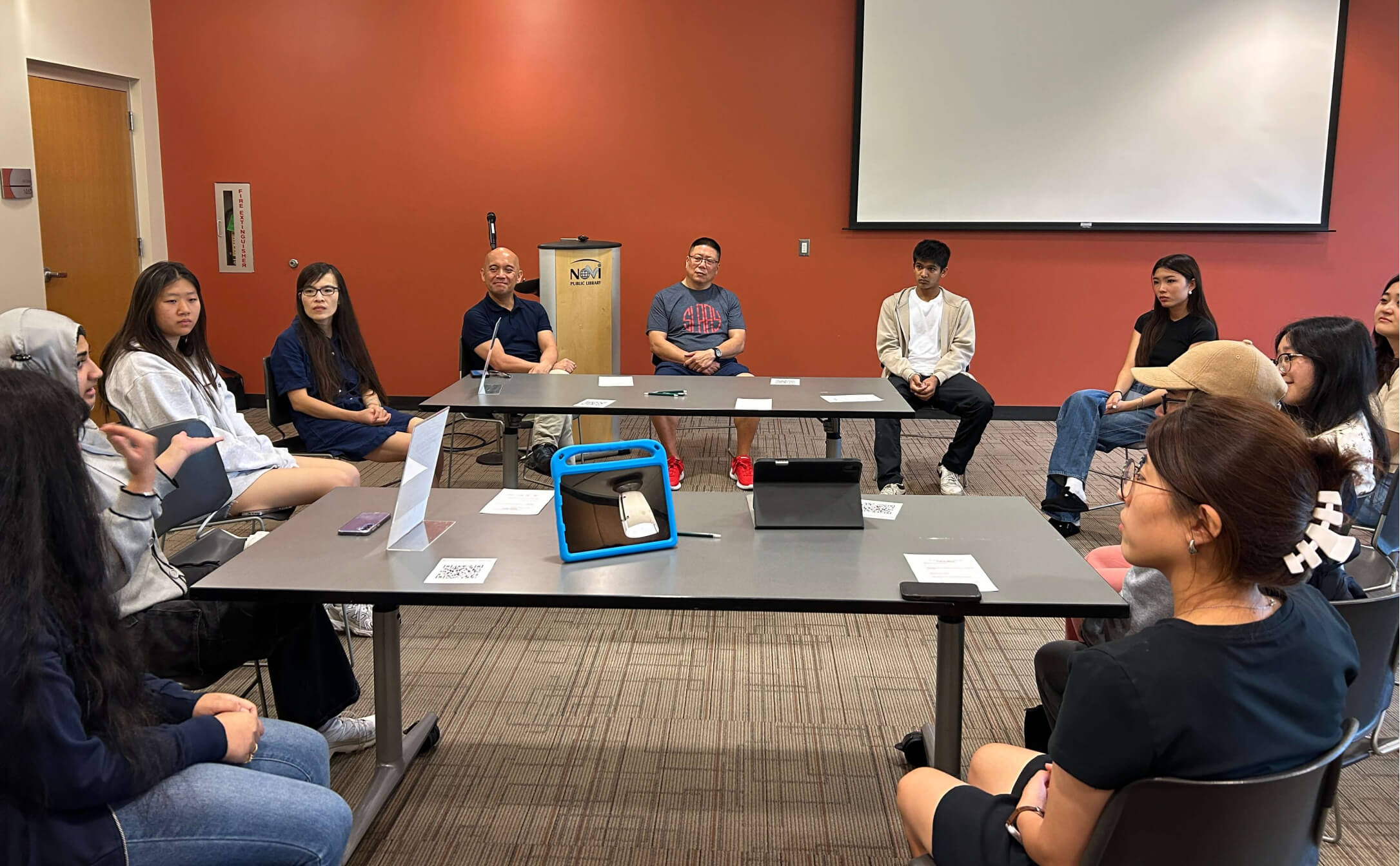 A group of people seated in a circle in a conference room with red walls, engaged in a discussion near a podium labeled Novi Public Library.