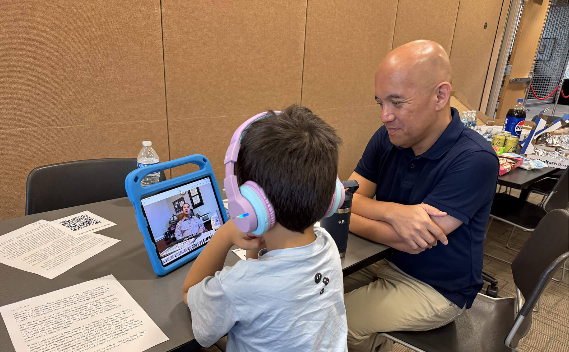 A child wearing pastel pink and blue headphones watches a video on a tablet while an adult man in a navy polo shirt sits across the table, watching attentively.