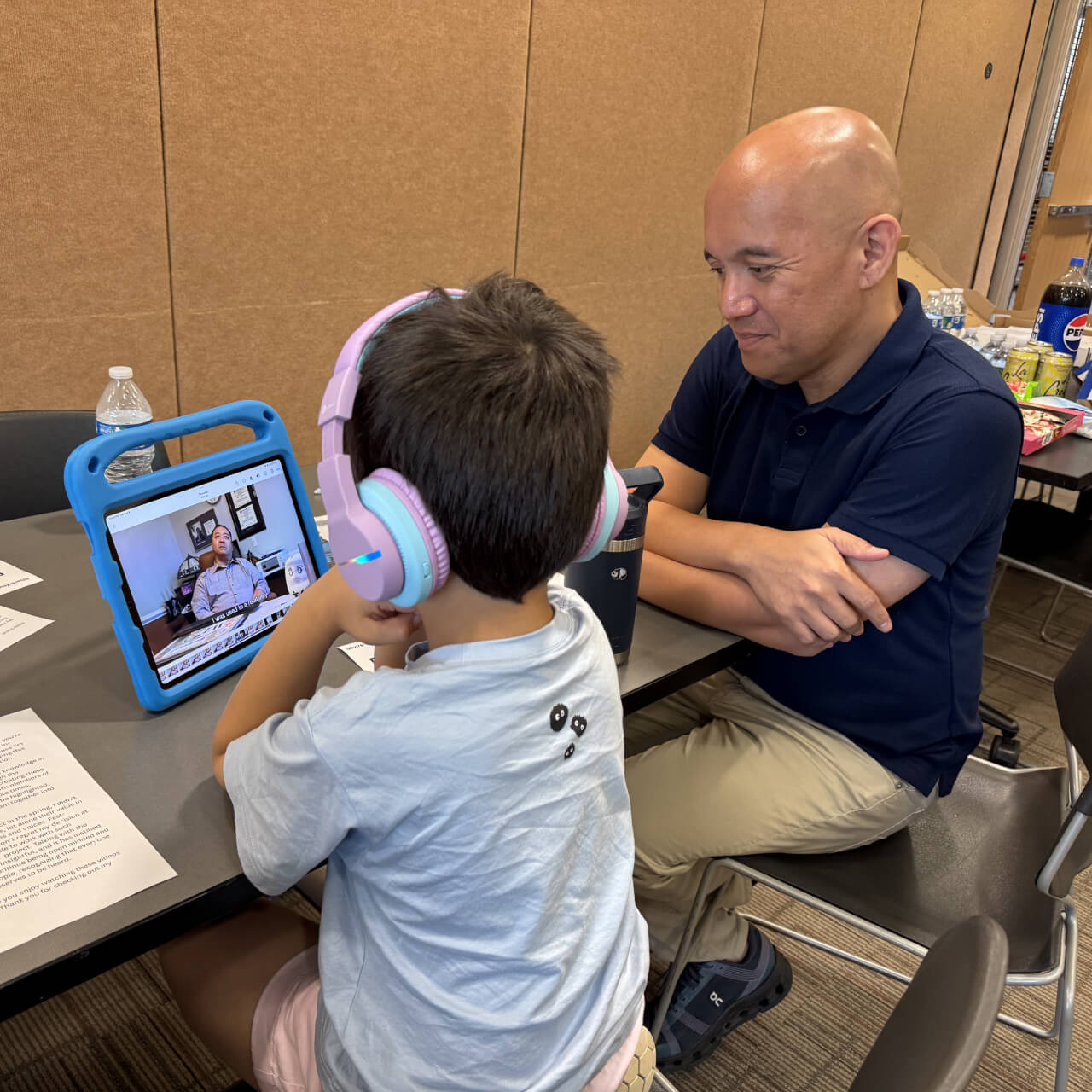 A child wearing pink and blue headphones watches a video on a tablet while an adult man in a navy shirt and khaki pants observes at a table.