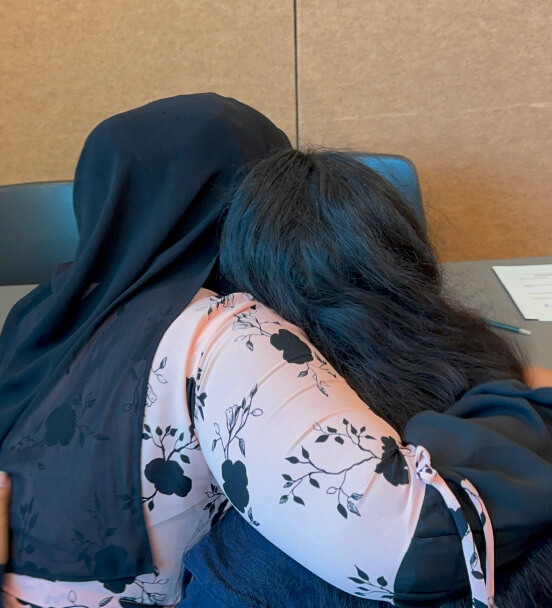 Two women embracing each other, one wearing a black hijab and the other with long dark hair and a floral patterned blouse.