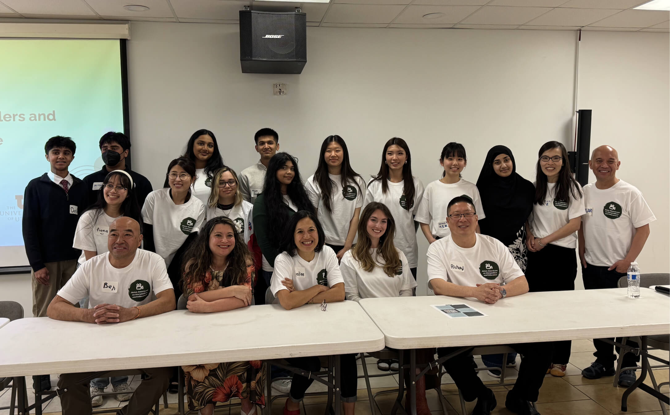 Group of diverse people in a room, some seated at tables, most wearing white event t-shirts, smiling.