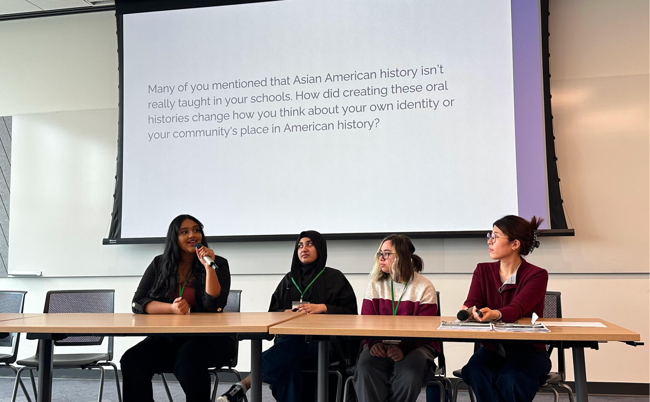 Panel of four diverse women seated with microphones under a screen showing text about Asian American history.