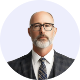 Middle-aged man with glasses, gray beard, wearing a dark checkered suit and patterned tie on a light background.