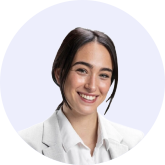 Smiling young woman with dark hair tied back wearing a white collared shirt and light gray blazer on a light background.