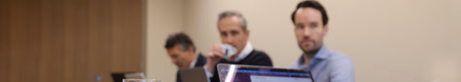 Three men sitting at a conference table with laptops, one drinking from a cup.
