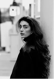 Black and white portrait of a woman with long wavy hair wearing a dark jacket, looking over her shoulder outdoors.