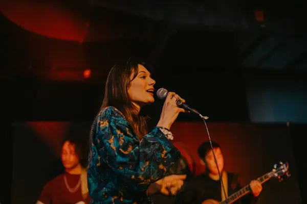 Female singer performing on stage with two musicians playing instruments in the background.