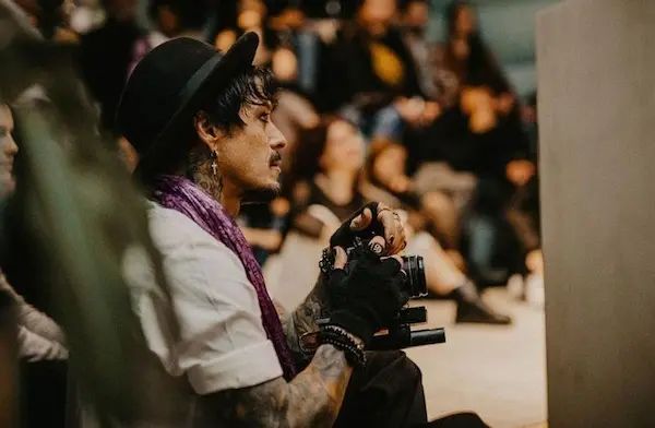 Man with tattoos and gloves holding a camera, wearing a hat and scarf, seated in an audience.