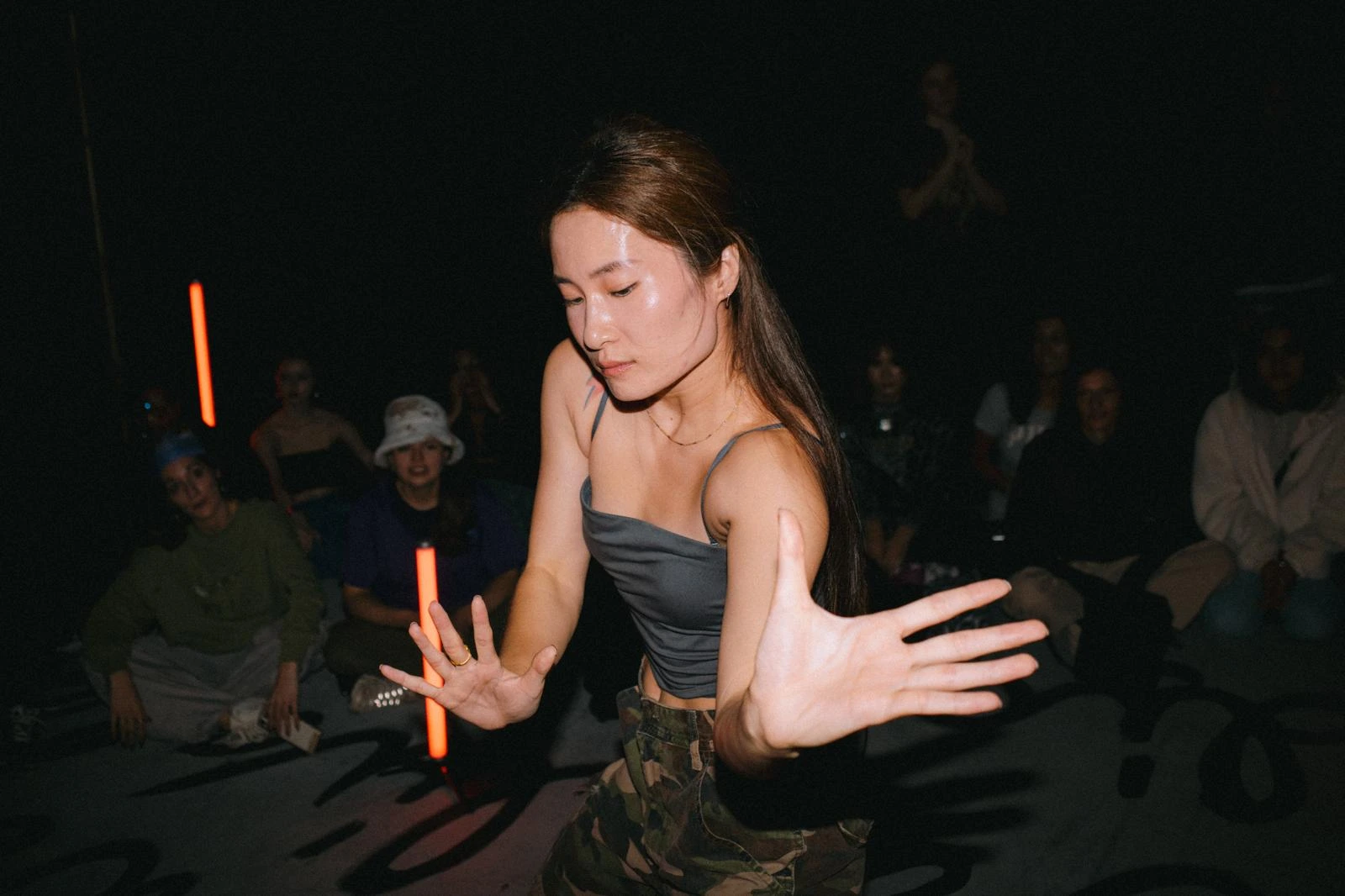 Young woman dancing with intense focus in a dimly lit room with seated audience in the background.