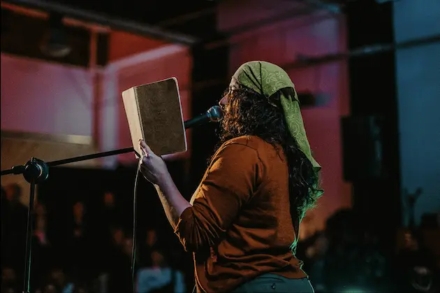 Person with curly hair wearing a green headscarf and brown shirt reading from a book into a microphone on stage.