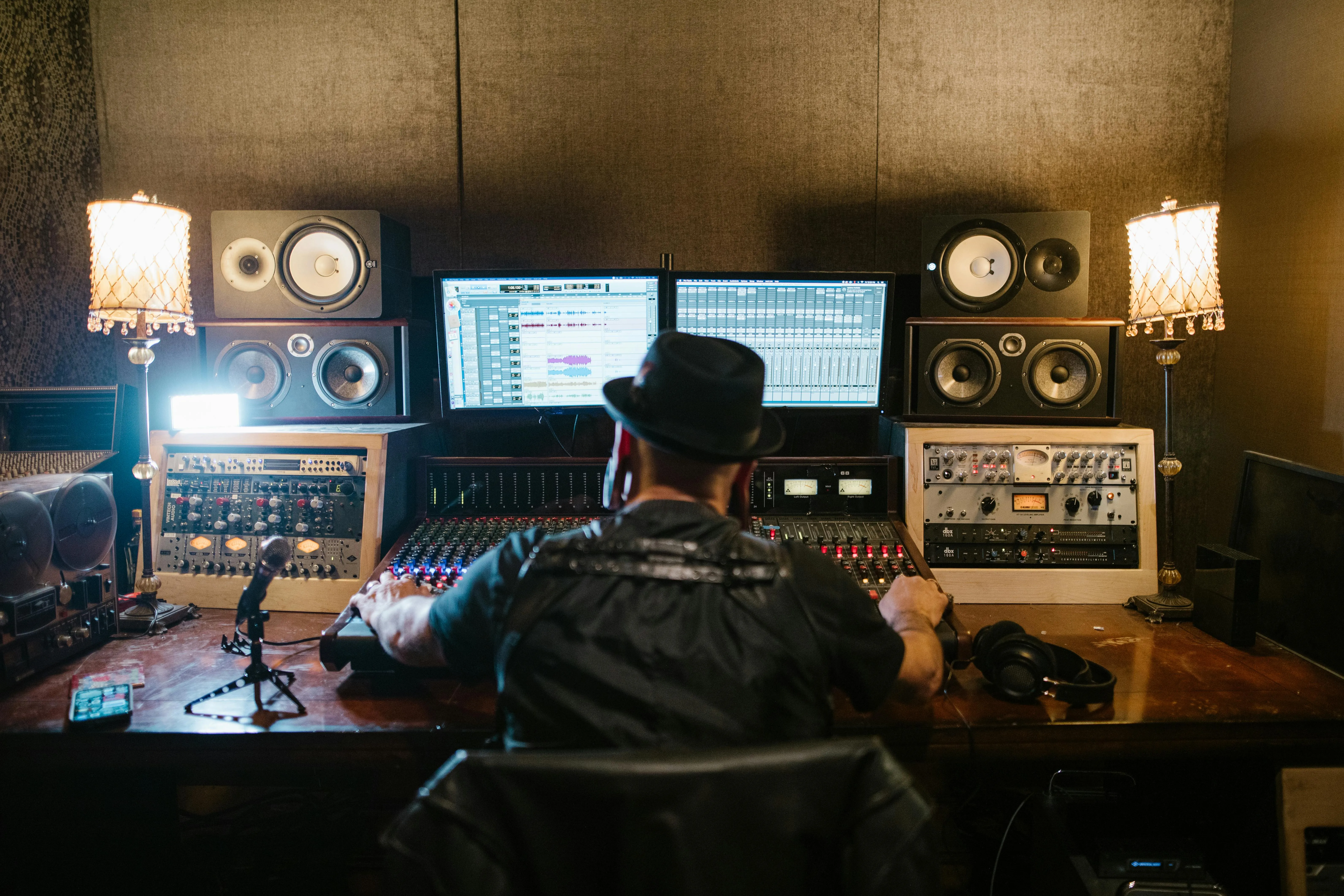 Person wearing a hat working at an audio mixing console with dual monitors and speakers in a recording studio.
