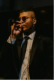 Man wearing sunglasses and a suit singing into a microphone on a dark stage.