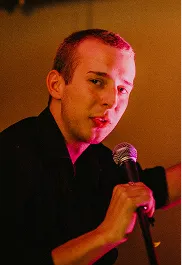 Young man with short hair holding a microphone and speaking or singing under warm, colorful lighting.
