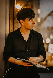 Young man in a black shirt holding a notebook and pen, looking thoughtfully to the side in a warmly lit indoor setting.