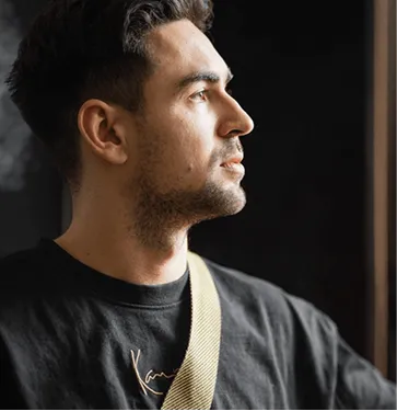 Young man looking thoughtfully to the right with a black shirt and beige strap over his shoulder.
