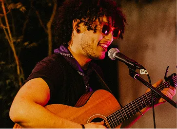 Musician with curly hair and sunglasses singing and playing acoustic guitar into a microphone.