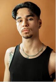 Young man with short dark hair, wearing a black tank top and gold chain against a brown background.