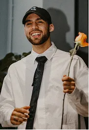 Smiling man wearing a black cap, white shirt, and black tie holding an orange rose.