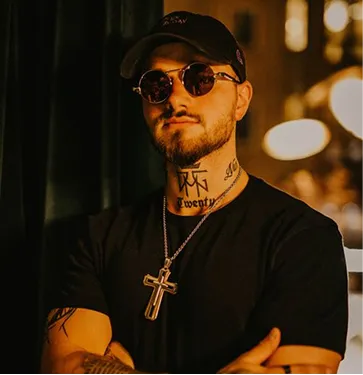Man wearing round sunglasses, black cap, and black shirt with cross necklace and visible neck tattoos.