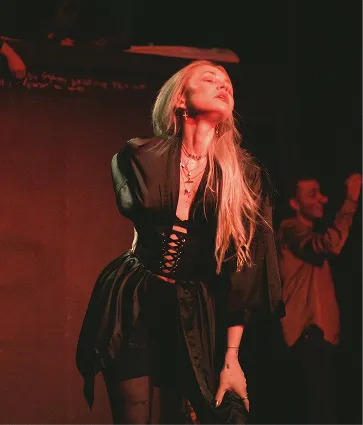 Woman with long blonde hair wearing a black dress, posing with eyes closed under warm lighting on stage.