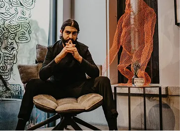 Man with long dark hair and beard sitting thoughtfully on a cushioned chair indoors, with an orange wireframe sculpture behind him.