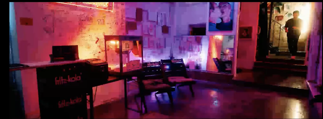 Dimly lit room with purple and orange lighting, two chairs, a table with a display case, and a person standing in a doorway.