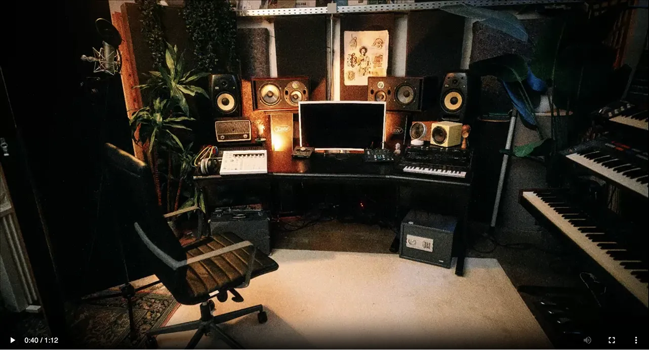 Home music studio with a computer monitor, multiple speakers, keyboards, and recording equipment on a desk surrounded by a swivel chair and plants.
