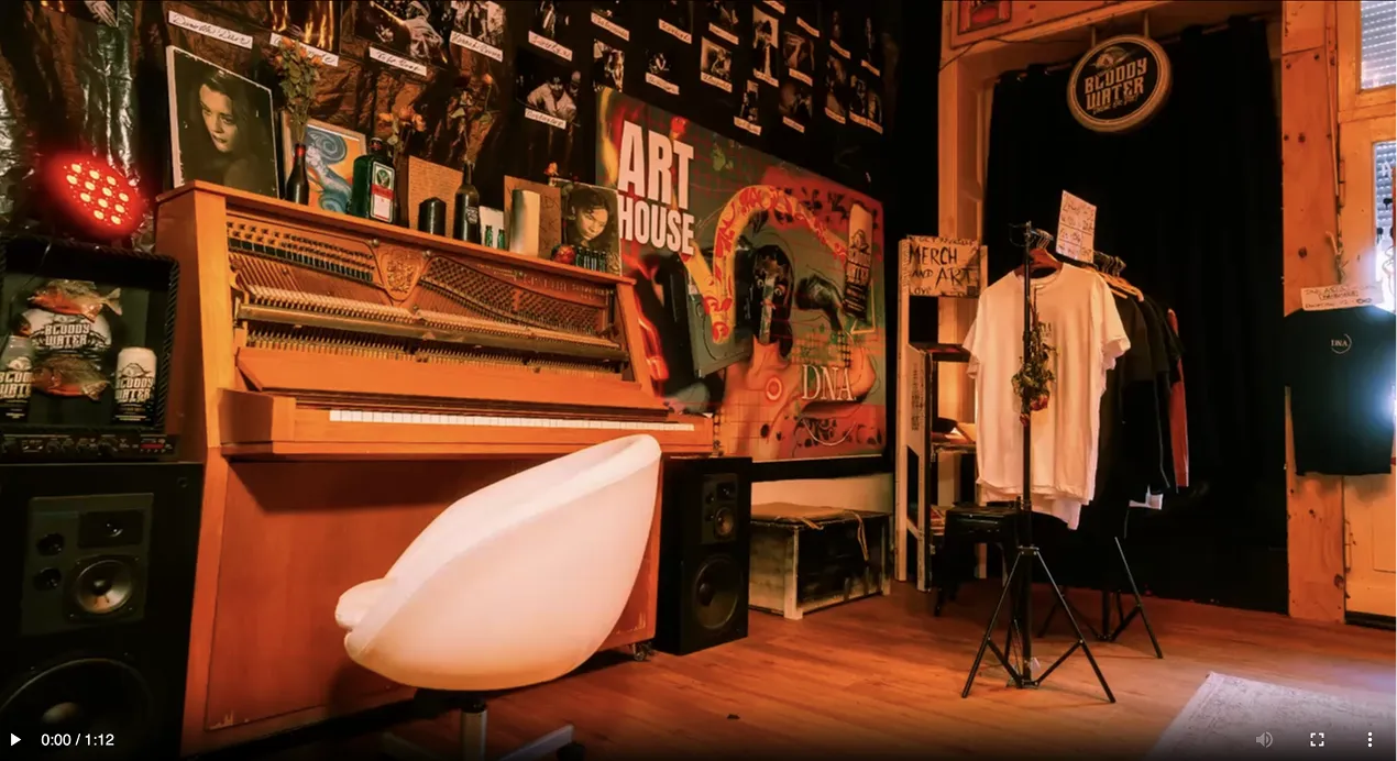 Cozy room with an open upright piano, a white swivel chair, framed photos, speakers, and a clothing rack with shirts for sale under warm lighting.