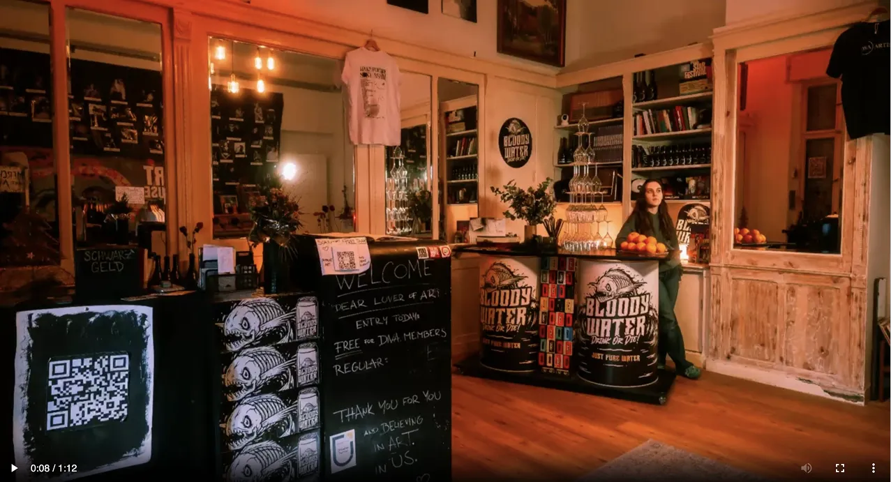 Interior of an art venue with a welcome sign, stacked Bloody Water drink barrels as counters, bookshelves, a woman standing behind the counter, and a warm ambient lighting.