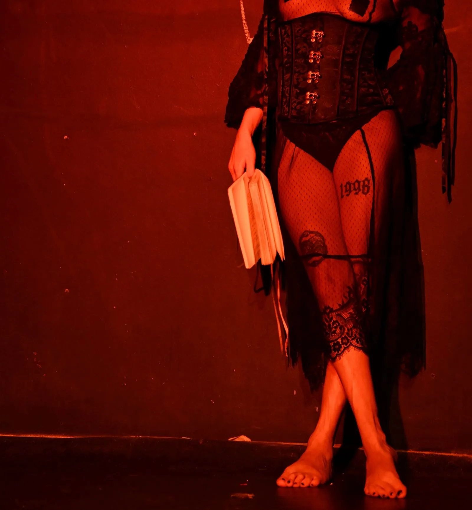 Person wearing black lace corset and fishnet stockings with a 1998 tattoo on thigh, holding an open book, standing barefoot against a dimly lit red wall.