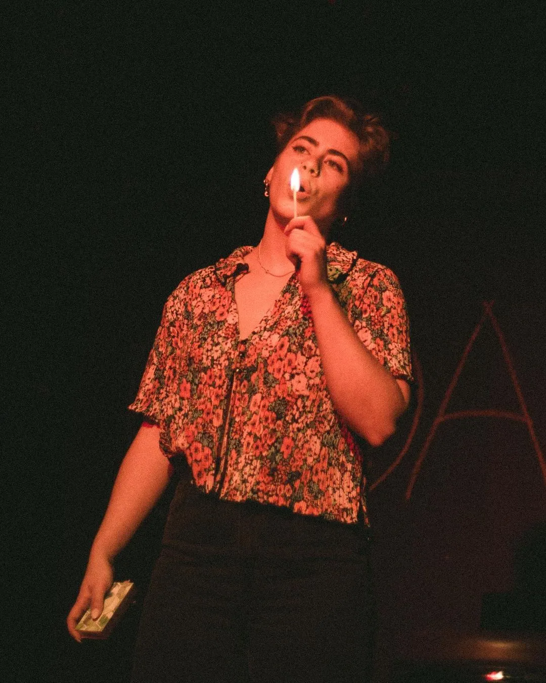Person wearing a floral shirt holding a lit matchstick close to their face against a dark background.