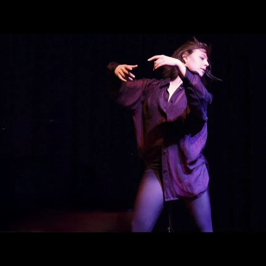 Woman in a loose purple shirt dancing with expressive arm movements against a dark background.