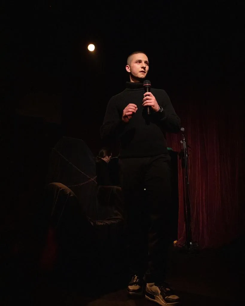 Man in black sweater holding a microphone and speaking on a dimly lit stage with dark red curtains in the background.