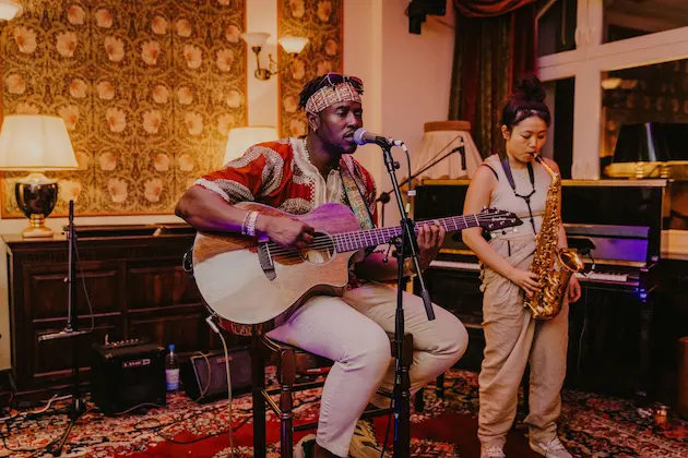 Man singing and playing an acoustic guitar while a woman plays saxophone in an indoor setting with vintage decor.