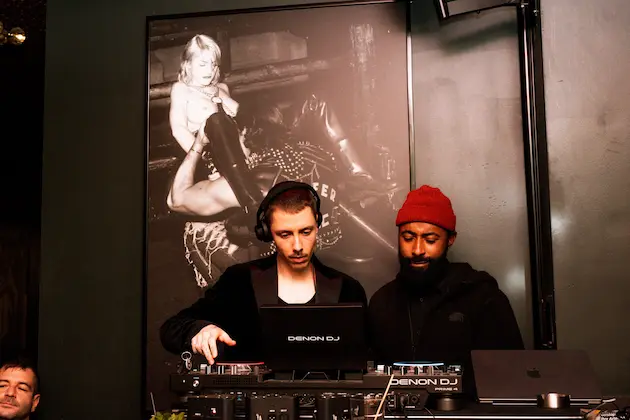 Two DJs performing with equipment in front of a large black-and-white poster of a child holding a boot.