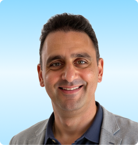 Smiling man with short dark hair wearing a gray blazer over a navy shirt against a light blue background.