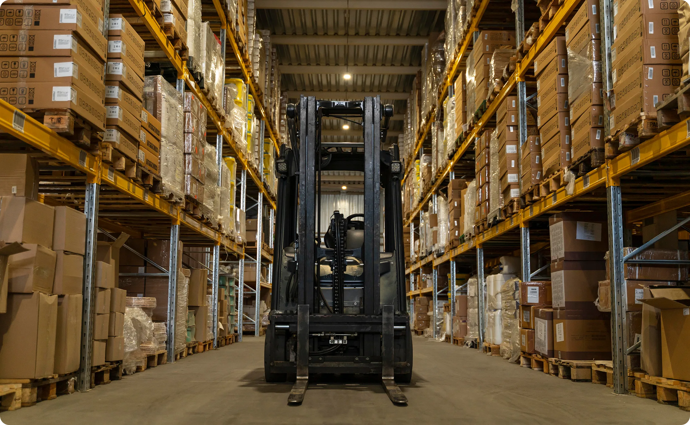 Two factory workers wearing yellow helmets and gray uniforms walking and discussing inside a spacious warehouse with stacked metal materials.