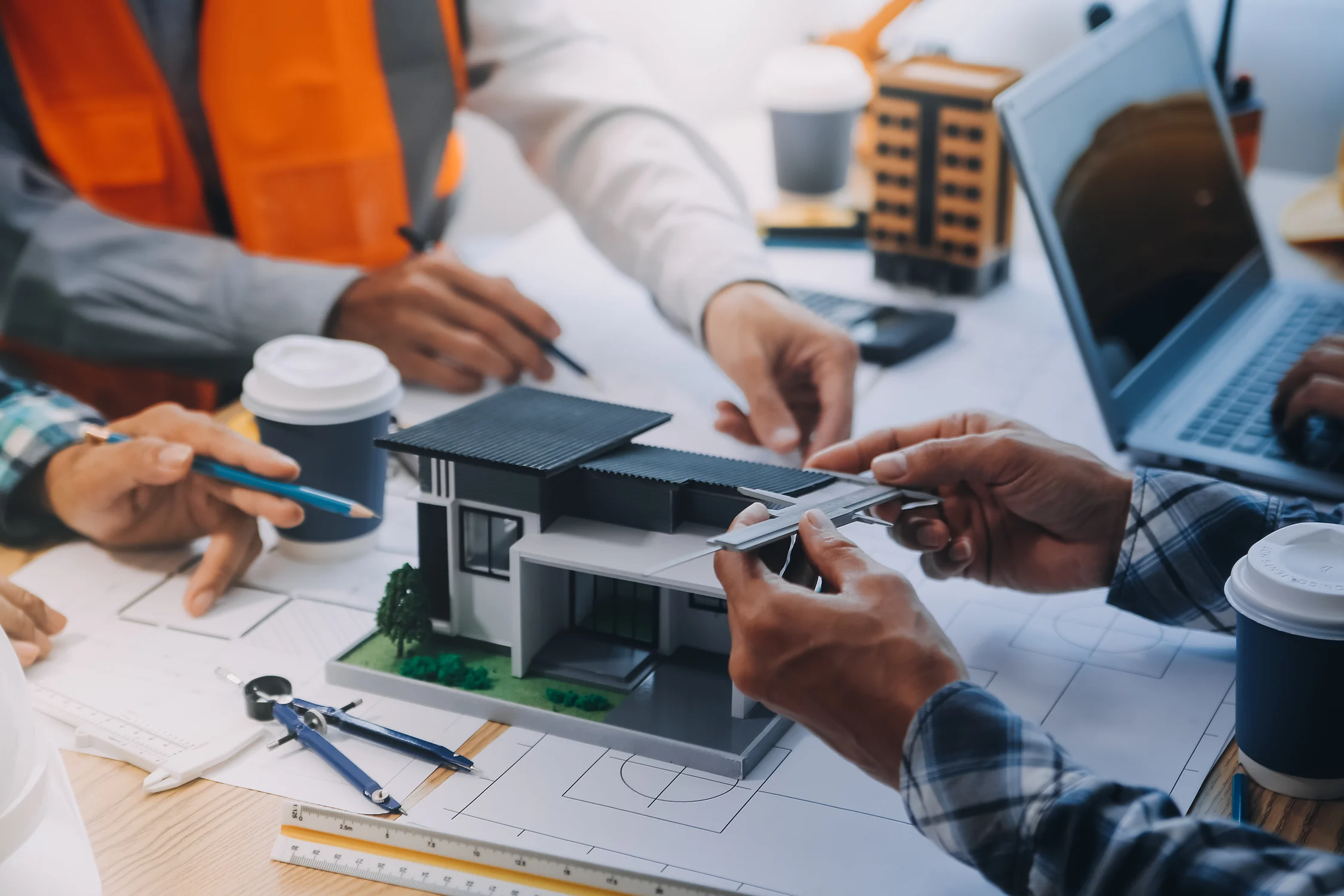 Architects or engineers discussing a modern house model with measuring tools on a desk with blueprints and laptops.
