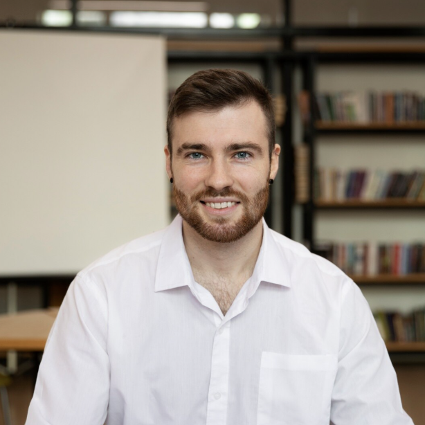 Smiling man with short brown hair, beard, and blue eyes wearing a white shirt, sitting indoors with bookshelves in the background.
