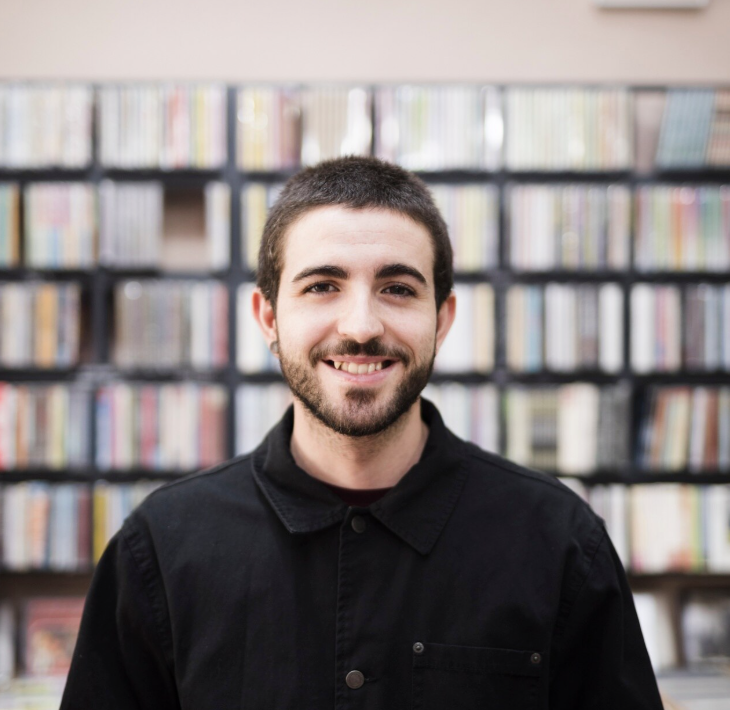 Smiling young man with short dark hair and beard wearing a black jacket, standing in front of a bookshelf.