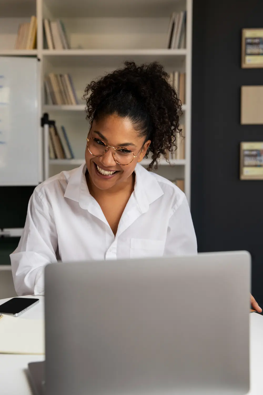 Smiling woman with curly hair and glasses using a laptop at a desk in an office.