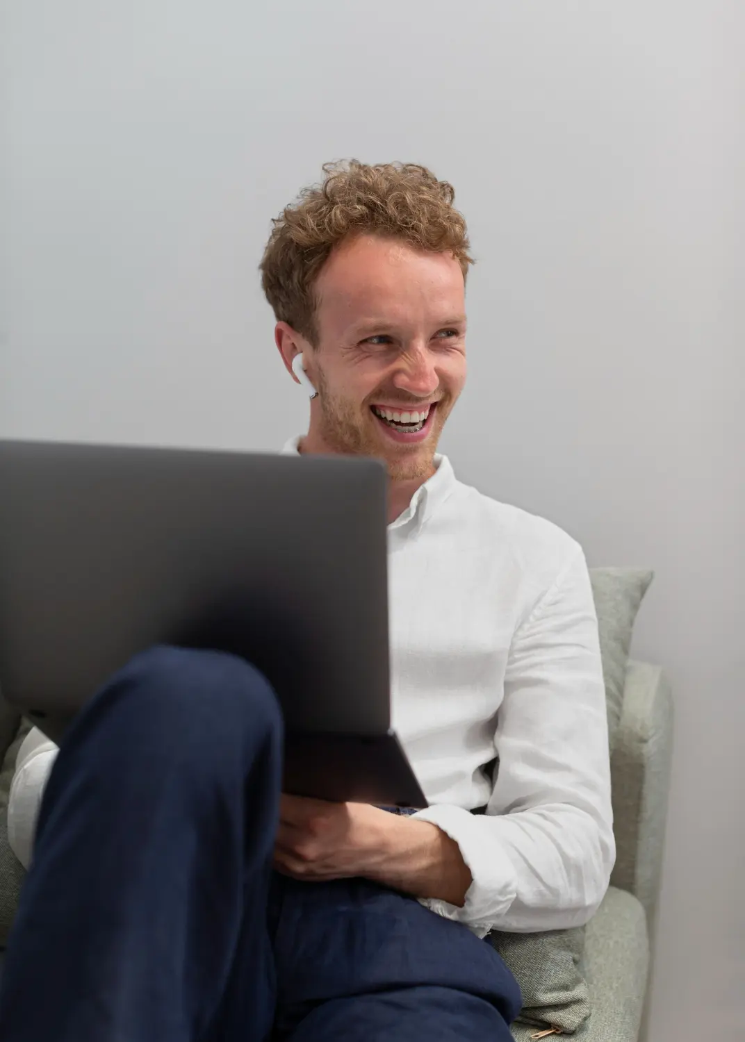 Smiling man with curly hair wearing a white shirt and wireless earbuds holding a laptop on his lap while sitting on a gray couch.