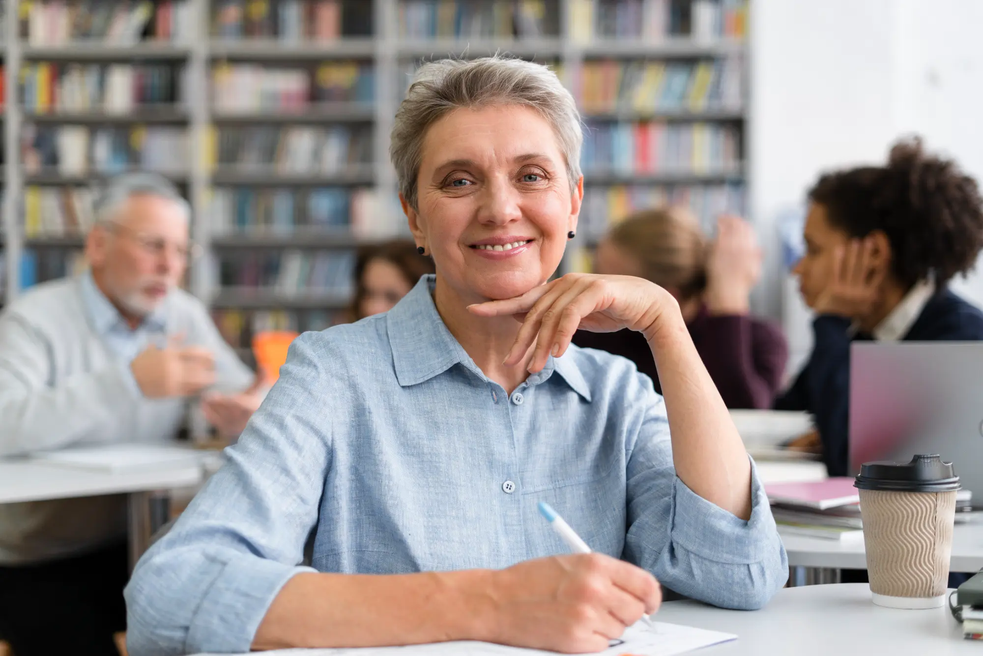Smiling senior woman sitting at a table in a library, writing on paper with a pen, with a coffee cup nearby and blurred people and bookshelves in the background.