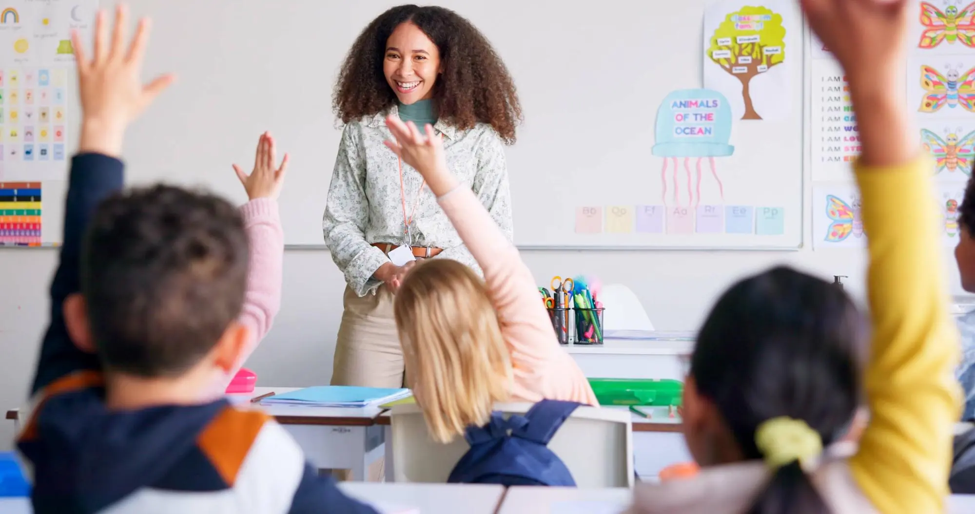 Smiling teacher standing in front of a whiteboard while students raise their hands in a classroom.
