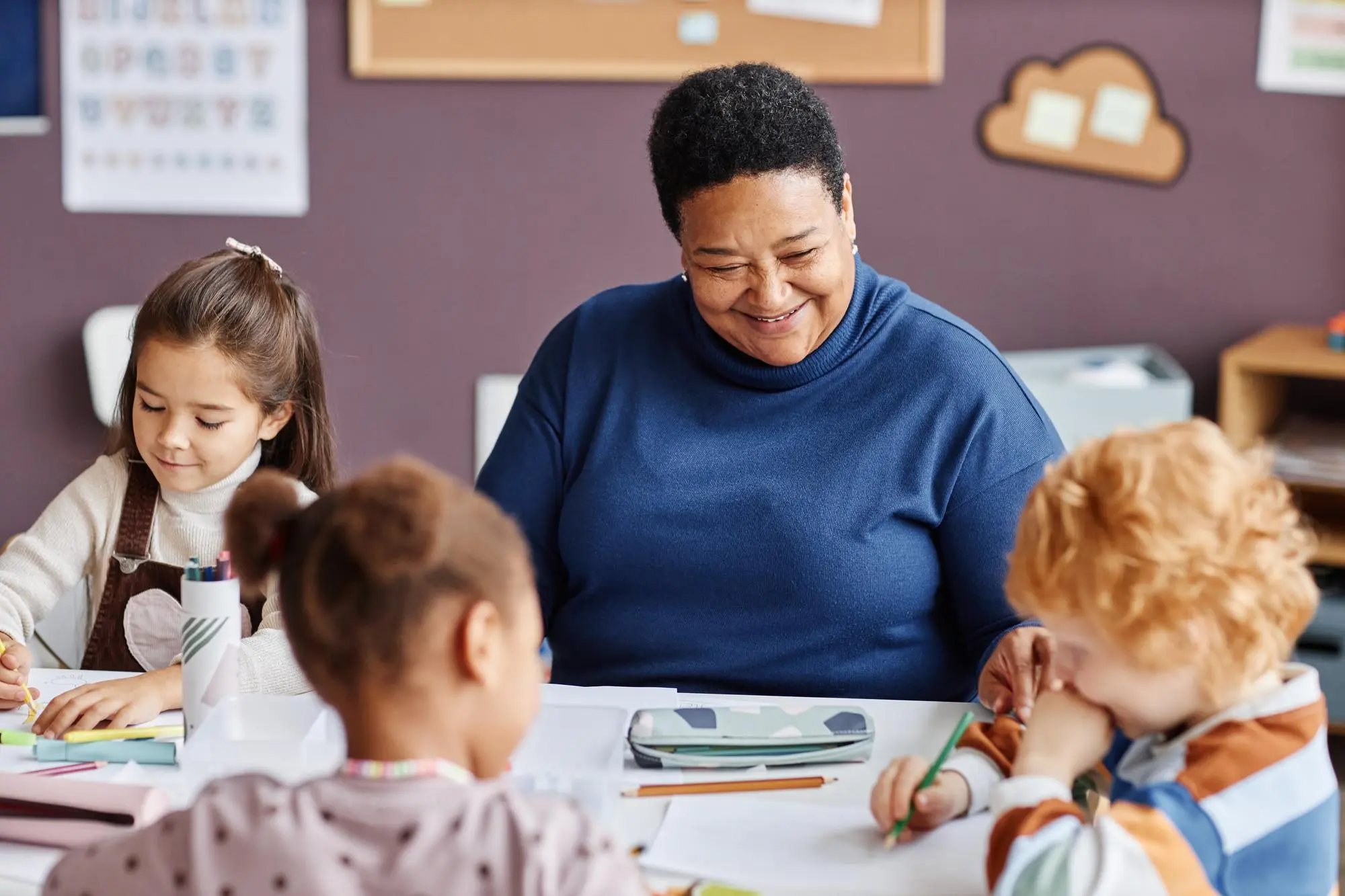 Smiling teacher sitting at a table with three young children who are drawing and coloring.