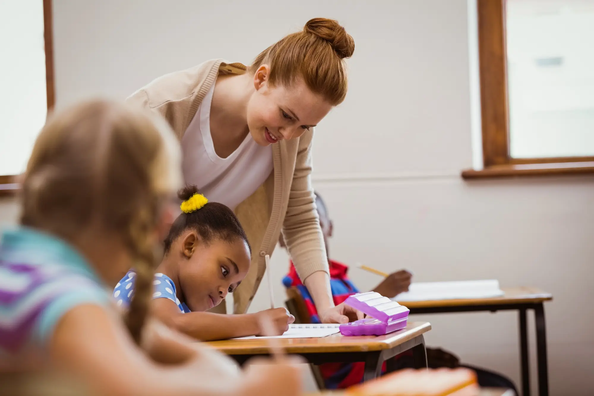 Teacher leaning over and assisting a young girl writing at her desk in a classroom.