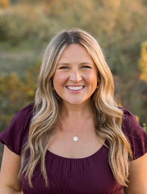 Smiling woman with long wavy blonde hair wearing a purple top and gold necklace outdoors.