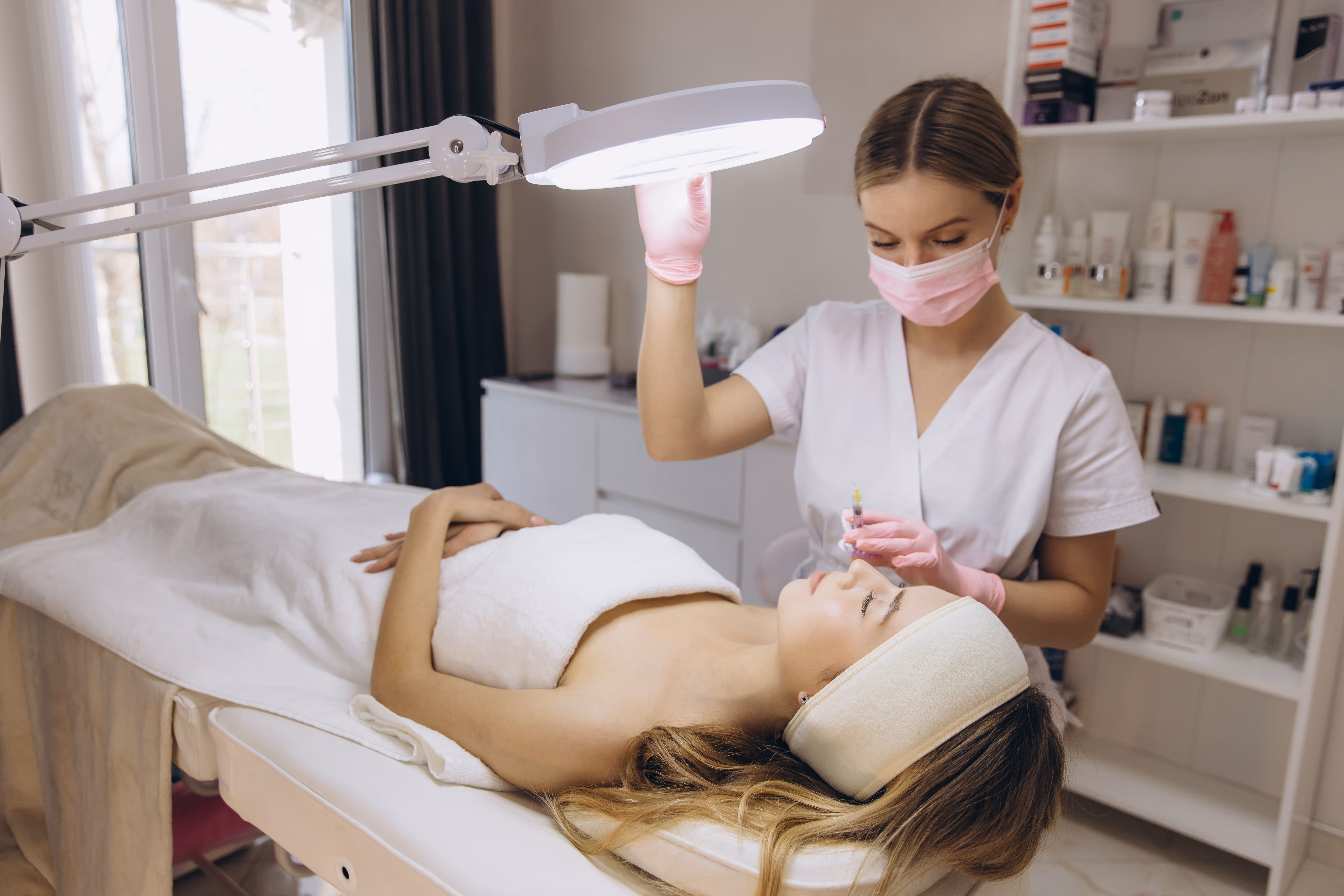 Beauty professional wearing pink gloves and mask preparing an injection for a woman lying on a treatment table in a spa room.