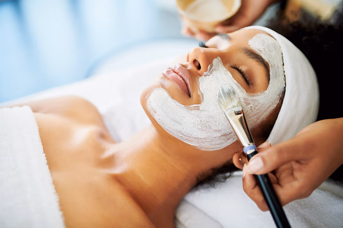 Woman receiving a facial treatment with a white mask applied by a brush at a spa.